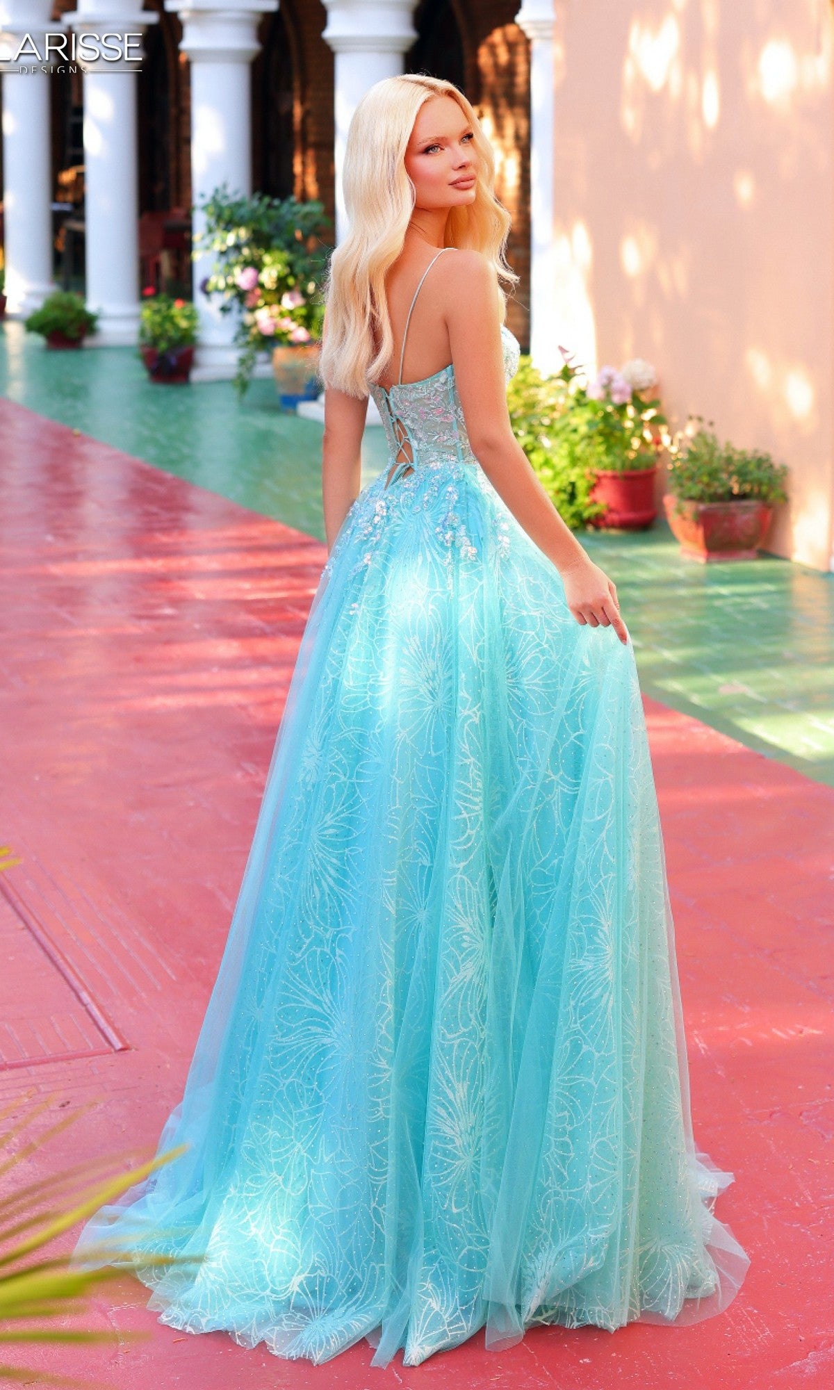 A woman with long blonde hair wears the Clarisse Designs 62310 long prom dress, featuring a light blue, floor-length corset bodice style, while standing outdoors on a red and green walkway near potted plants.